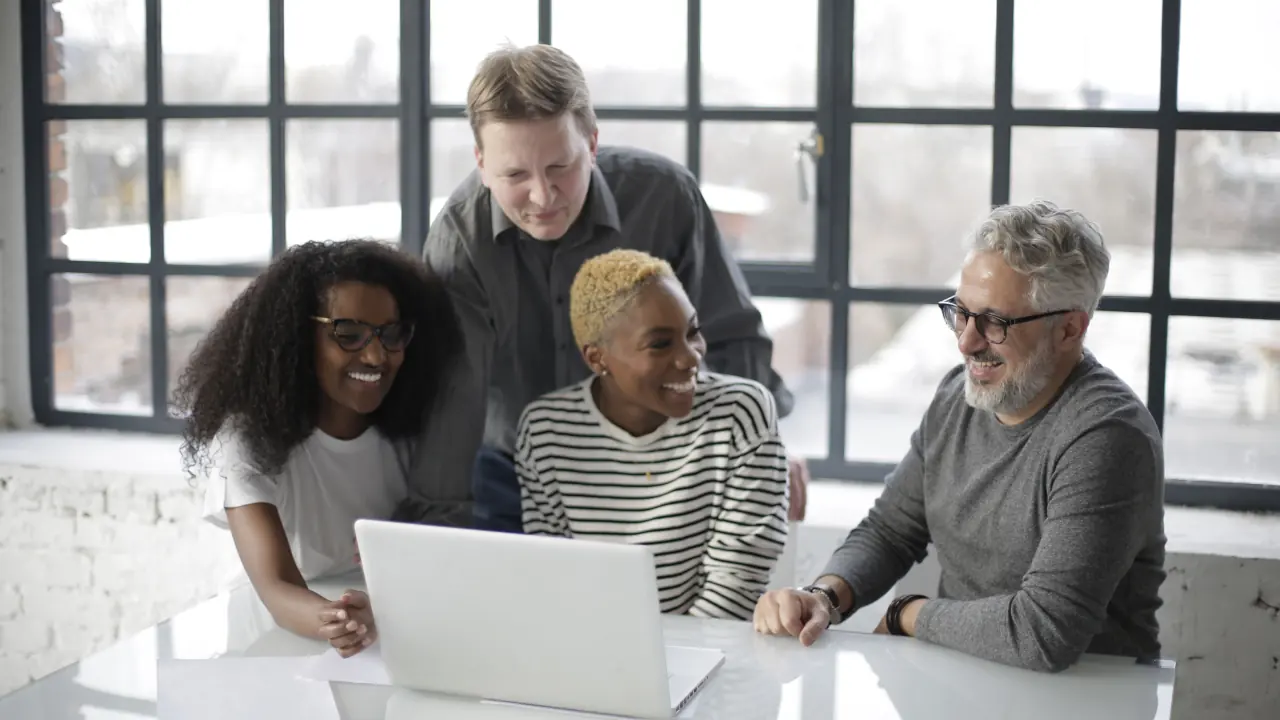 Four colleagues gather round a laptop at a white table in front of a large window